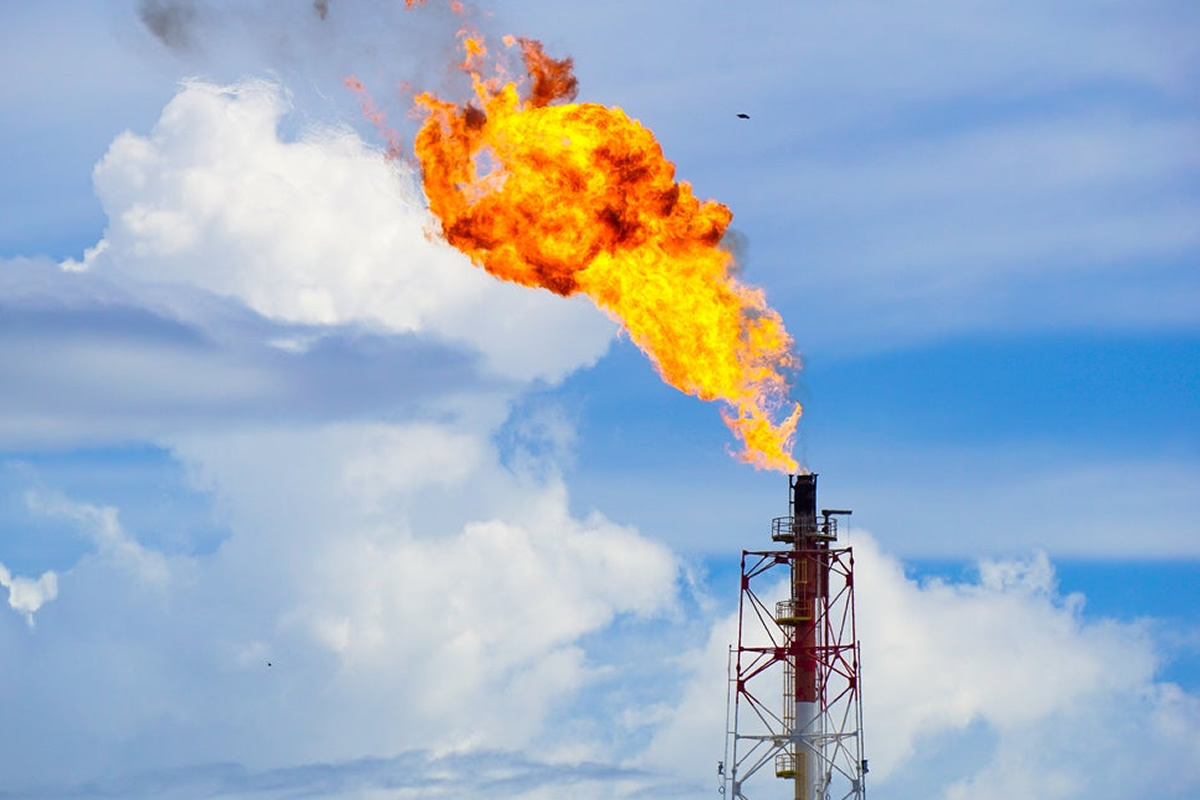 Industrial flare stack emitting a large orange flame into a blue sky with white clouds, at a processing facility