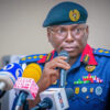 Military/paramilitary officer in a blue uniform and teal beret speaks into a microphone at a press conference, surrounded by multiple news microphones and reporters.