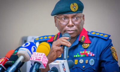 Military/paramilitary officer in a blue uniform and teal beret speaks into a microphone at a press conference, surrounded by multiple news microphones and reporters.
