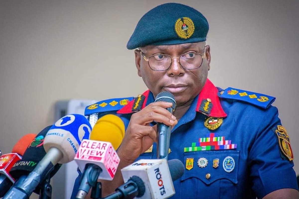 Military/paramilitary officer in a blue uniform and teal beret speaks into a microphone at a press conference, surrounded by multiple news microphones and reporters.
