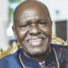 Smiling man in a dark blue traditional outfit with a gold chain, seated on an ornate chair in a bright setting.