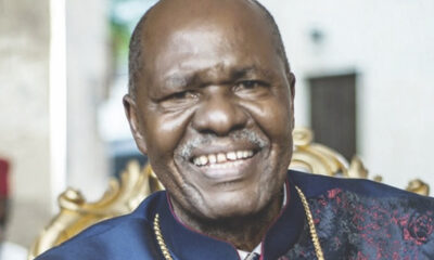 Smiling man in a dark blue traditional outfit with a gold chain, seated on an ornate chair in a bright setting.