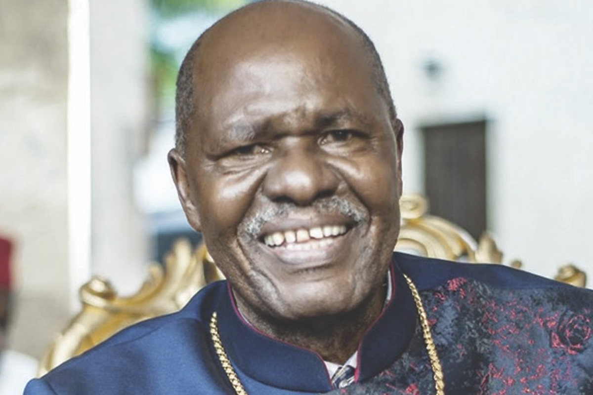 Smiling man in a dark blue traditional outfit with a gold chain, seated on an ornate chair in a bright setting.