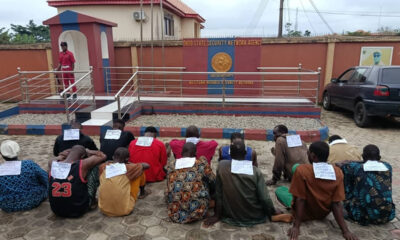 Group of men seated on the ground with papers on their backs, facing a security facility entrance with a guard watching nearby.
