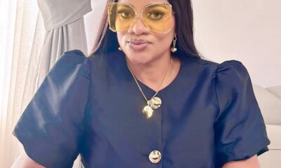 Woman seated indoors wearing oversized yellow sunglasses, navy puff-sleeve blouse, and gold jewelry.