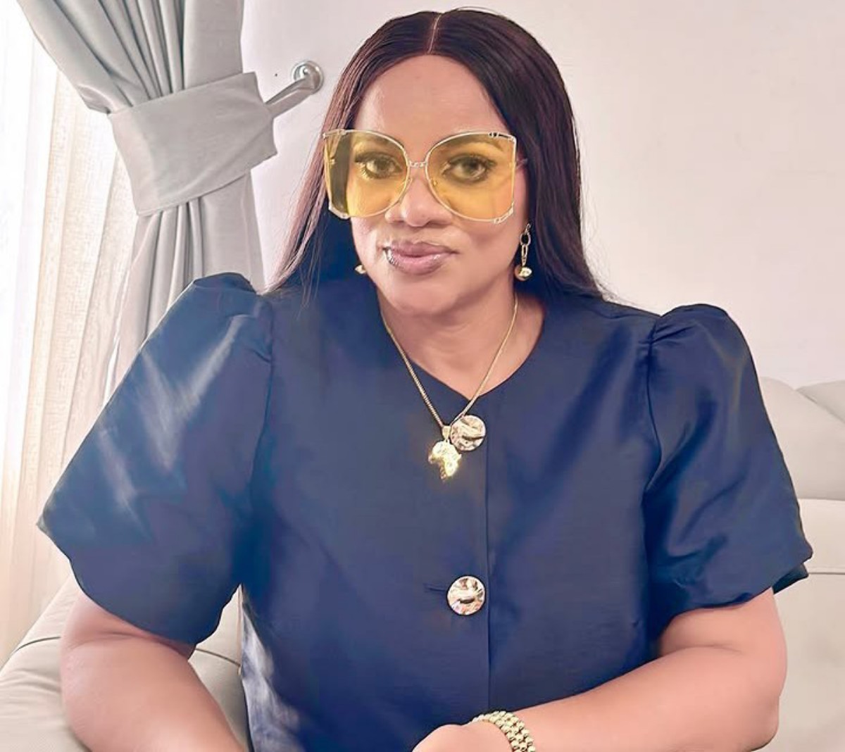 Woman seated indoors wearing oversized yellow sunglasses, navy puff-sleeve blouse, and gold jewelry.