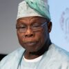 African man in light blue surgical scrubs and cap, wearing glasses, looking to the side with a thoughtful expression.