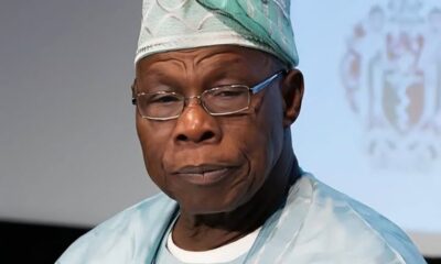 African man in light blue surgical scrubs and cap, wearing glasses, looking to the side with a thoughtful expression.