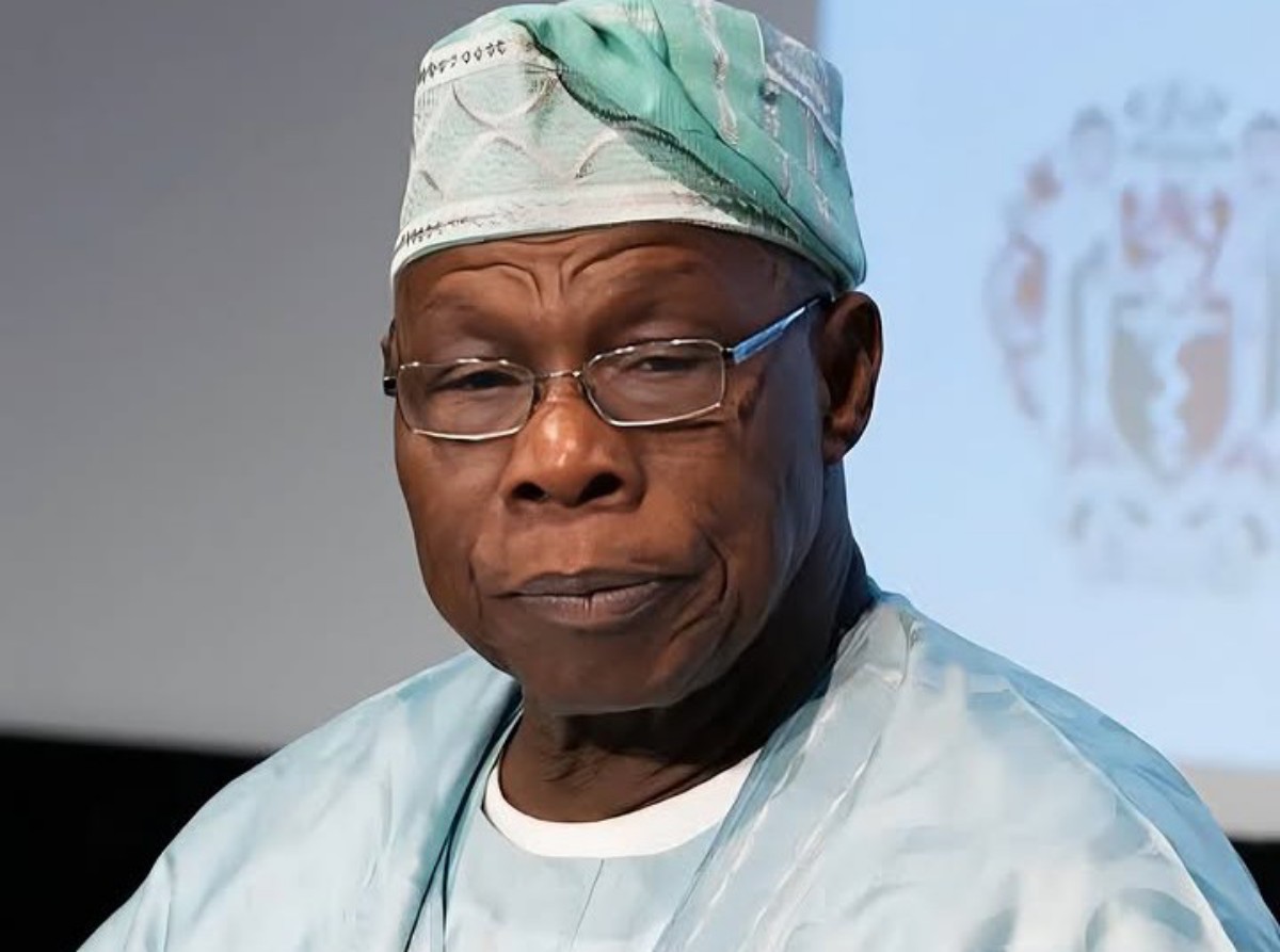 African man in light blue surgical scrubs and cap, wearing glasses, looking to the side with a thoughtful expression.