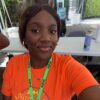 Smiling young woman wearing an orange shirt and a green ADMA lanyard, taking a selfie outdoors under a canopy.