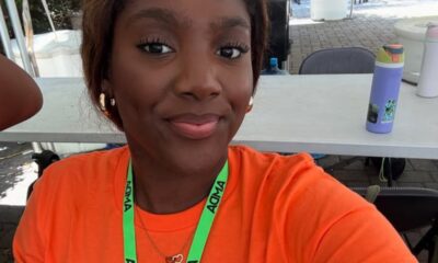 Smiling young woman wearing an orange shirt and a green ADMA lanyard, taking a selfie outdoors under a canopy.