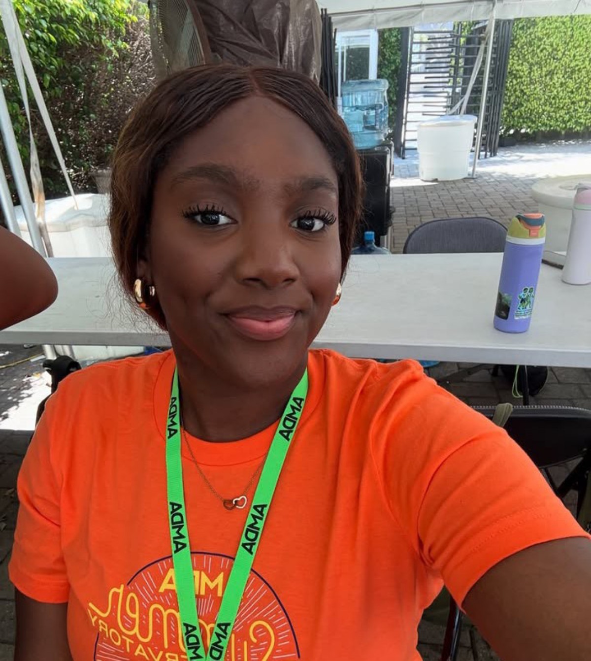Smiling young woman wearing an orange shirt and a green ADMA lanyard, taking a selfie outdoors under a canopy.
