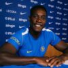 Chelsea FC player in a blue jersey smiles at the camera during a press conference with sponsor logos in the background.