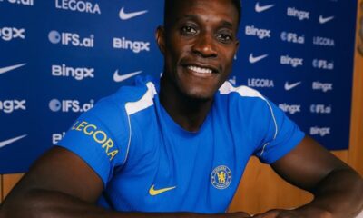Chelsea FC player in a blue jersey smiles at the camera during a press conference with sponsor logos in the background.