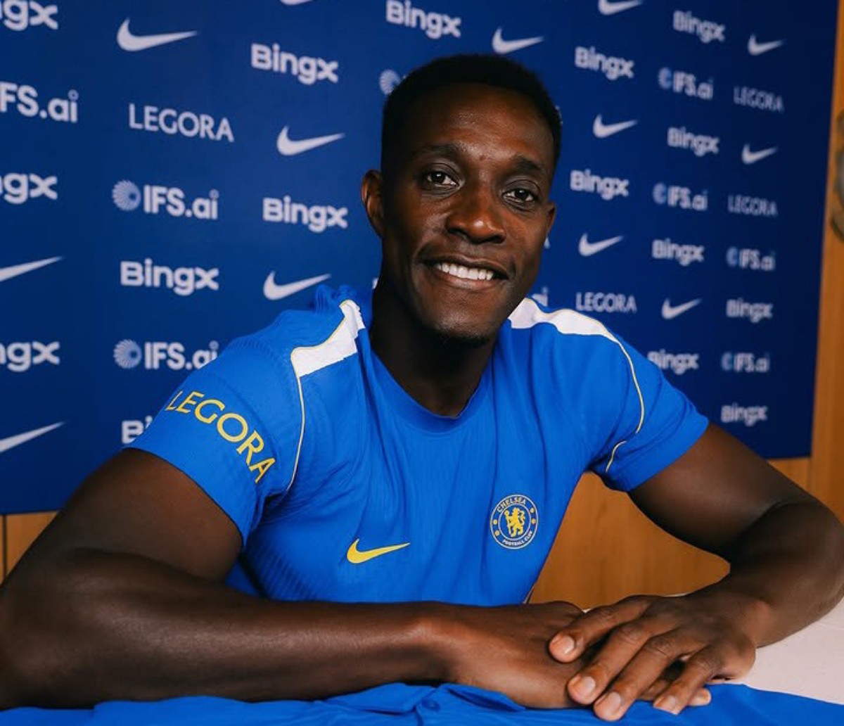 Chelsea FC player in a blue jersey smiles at the camera during a press conference with sponsor logos in the background.