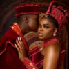 Couple in red ceremonial attire; man leans in to kiss the woman's temple as she gazes at the camera in a beaded headdress and dress.