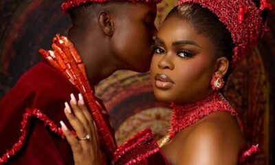 Couple in red ceremonial attire; man leans in to kiss the woman's temple as she gazes at the camera in a beaded headdress and dress.