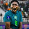 Brazilian soccer player in teal jersey shouting with mouth wide open, stadium crowd in the background