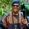 Man wearing a colorful traditional robe and hat, smiling, holding a carved staff in a lush garden setting.
