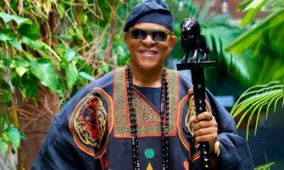 Man wearing a colorful traditional robe and hat, smiling, holding a carved staff in a lush garden setting.