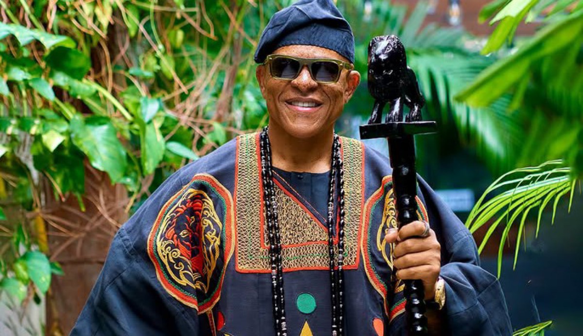 Man wearing a colorful traditional robe and hat, smiling, holding a carved staff in a lush garden setting.