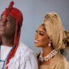 Couple in traditional Nigerian wedding attire: man in red beaded headdress and white outfit, woman in gold gele and pearl jewelry smiling side by side.
