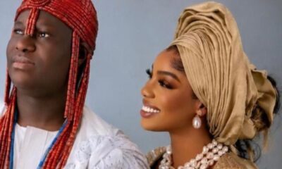 Couple in traditional Nigerian wedding attire: man in red beaded headdress and white outfit, woman in gold gele and pearl jewelry smiling side by side.
