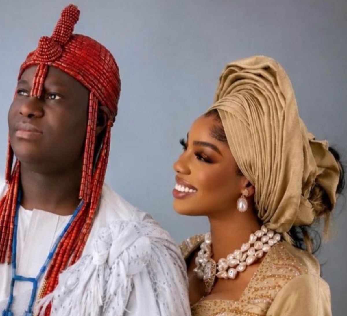 Couple in traditional Nigerian wedding attire: man in red beaded headdress and white outfit, woman in gold gele and pearl jewelry smiling side by side.