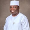 Smiling man in a white traditional outfit and white cap, posed against a neutral brown studio backdrop.