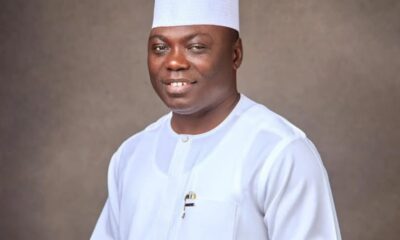 Smiling man in a white traditional outfit and white cap, posed against a neutral brown studio backdrop.