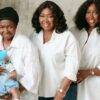 Three women standing together in white shirts, one holding a baby whose face is covered with a blue heart sticker.