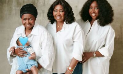 Three women standing together in white shirts, one holding a baby whose face is covered with a blue heart sticker.