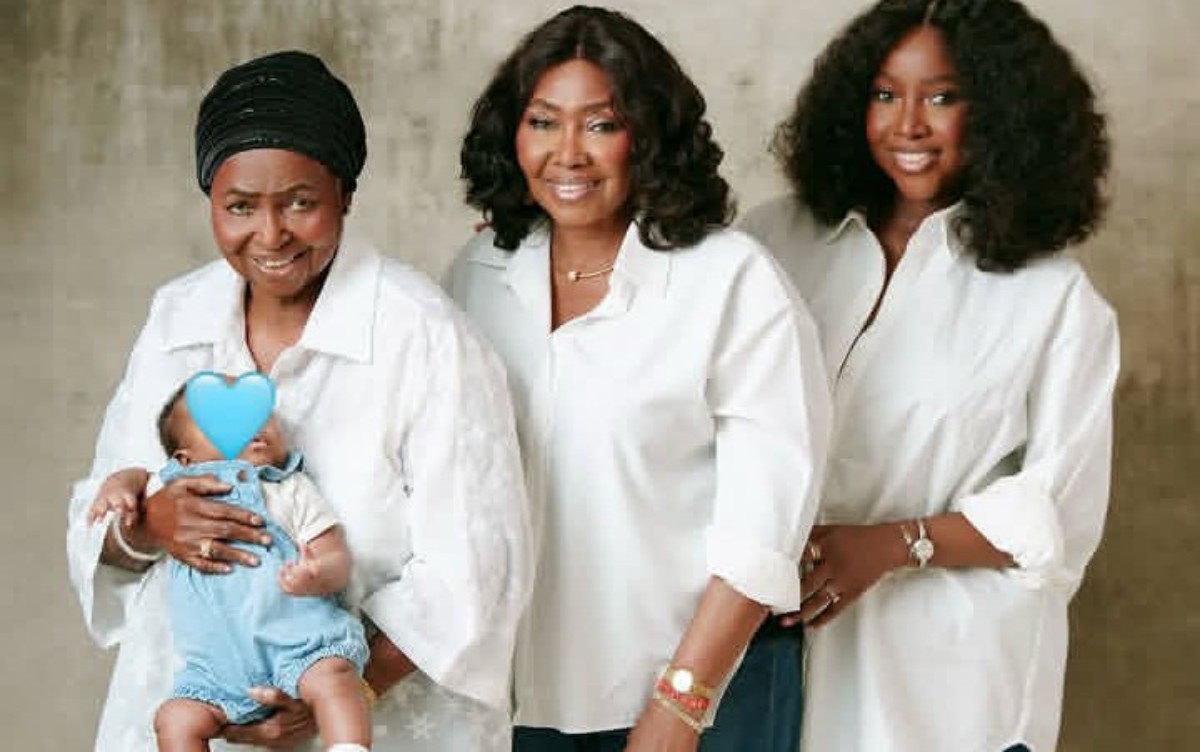 Three women standing together in white shirts, one holding a baby whose face is covered with a blue heart sticker.