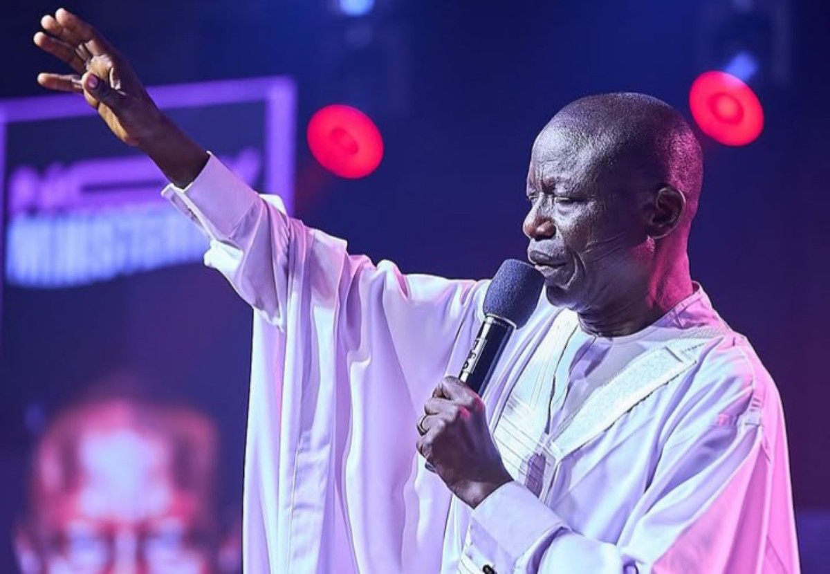 Male performer in a white robe sings into a microphone on stage, one arm raised in the air as he performs to the audience.