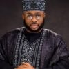 Portrait of a man wearing a black and silver patterned agbada with a matching cap and glasses, looking at the camera.