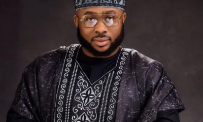 Portrait of a man wearing a black and silver patterned agbada with a matching cap and glasses, looking at the camera.