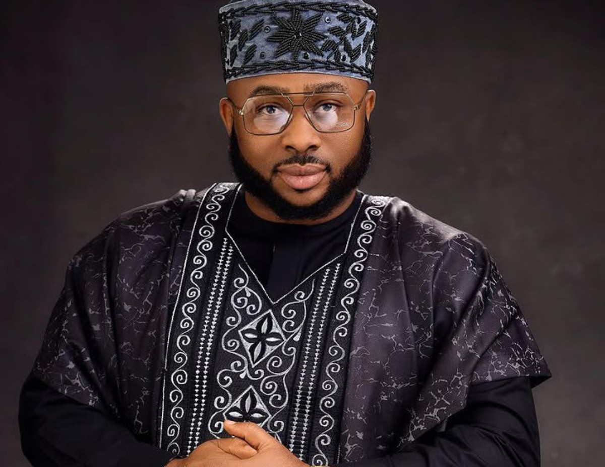 Portrait of a man wearing a black and silver patterned agbada with a matching cap and glasses, looking at the camera.