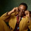 Man in a mustard suit sits on a vintage chair, adjusting sunglasses.