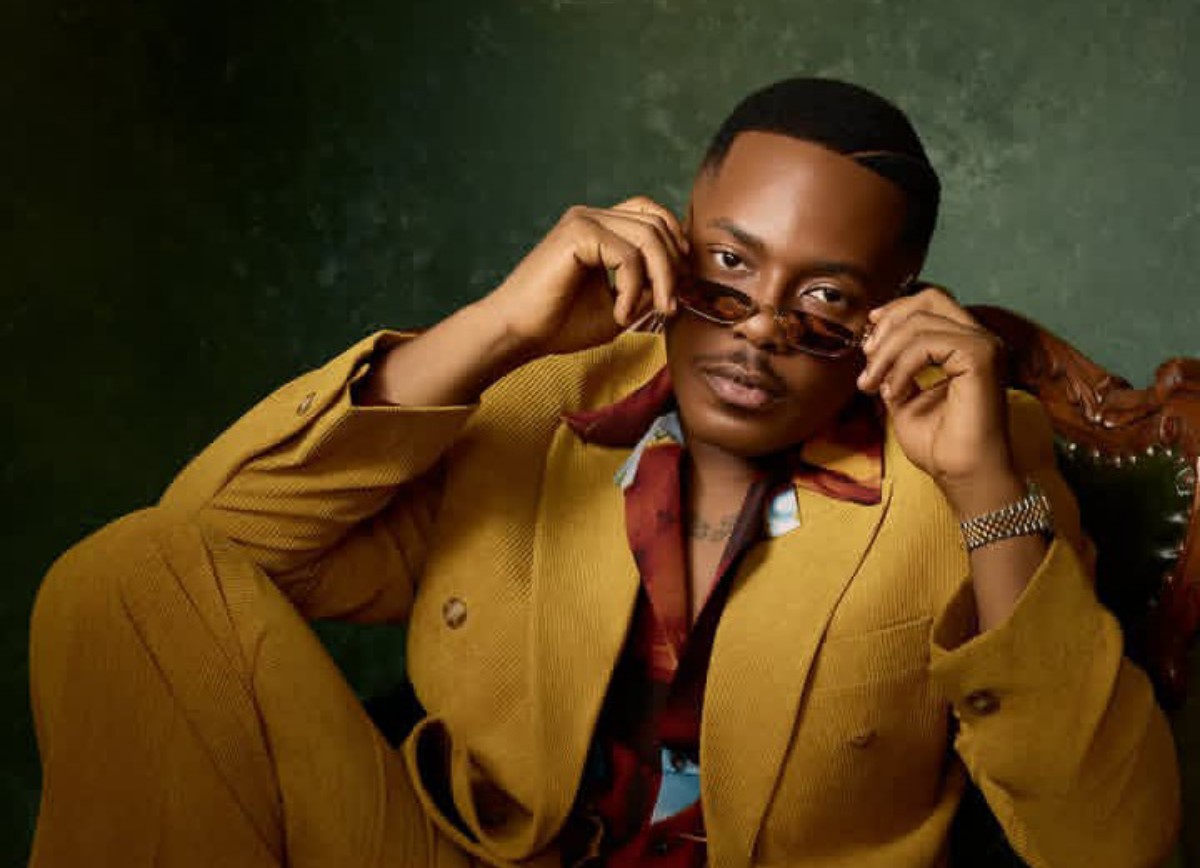 Man in a mustard suit sits on a vintage chair, adjusting sunglasses.
