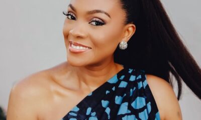 Smiling woman with a high ponytail and one-shoulder blue patterned dress, wearing sparkling stud earrings.