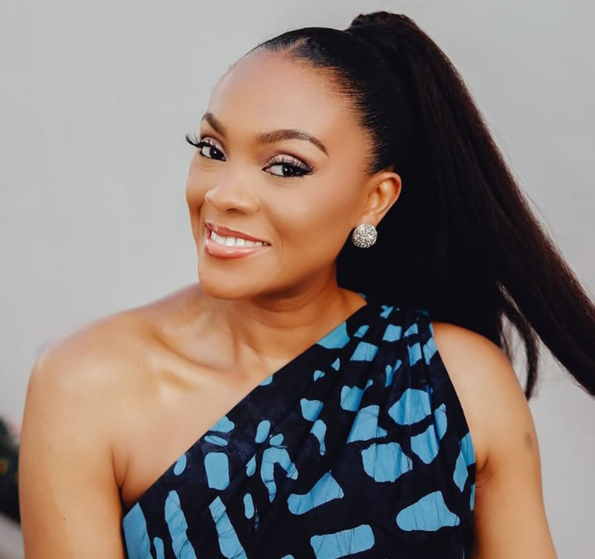 Smiling woman with a high ponytail and one-shoulder blue patterned dress, wearing sparkling stud earrings.