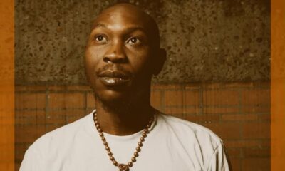 Portrait of a man wearing a beaded necklace, looking upward against a brick wall backdrop with an orange frame.