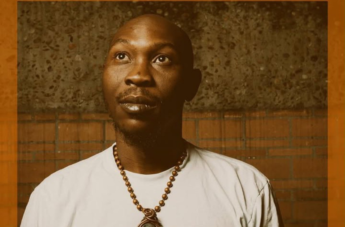 Portrait of a man wearing a beaded necklace, looking upward against a brick wall backdrop with an orange frame.