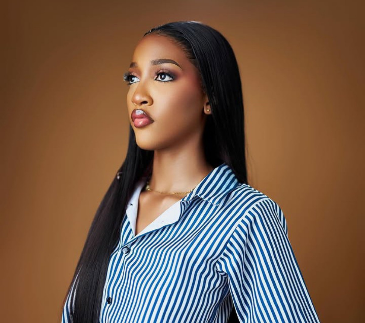 Woman with long black hair wearing a blue-and-white striped shirt, looking upward against a brown background.
