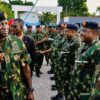 Senior army officer saluting as a line of soldiers in camouflage uniforms stand at attention outdoors, badges visible on uniforms.