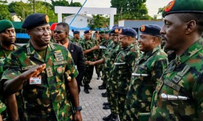 Senior army officer saluting as a line of soldiers in camouflage uniforms stand at attention outdoors, badges visible on uniforms.