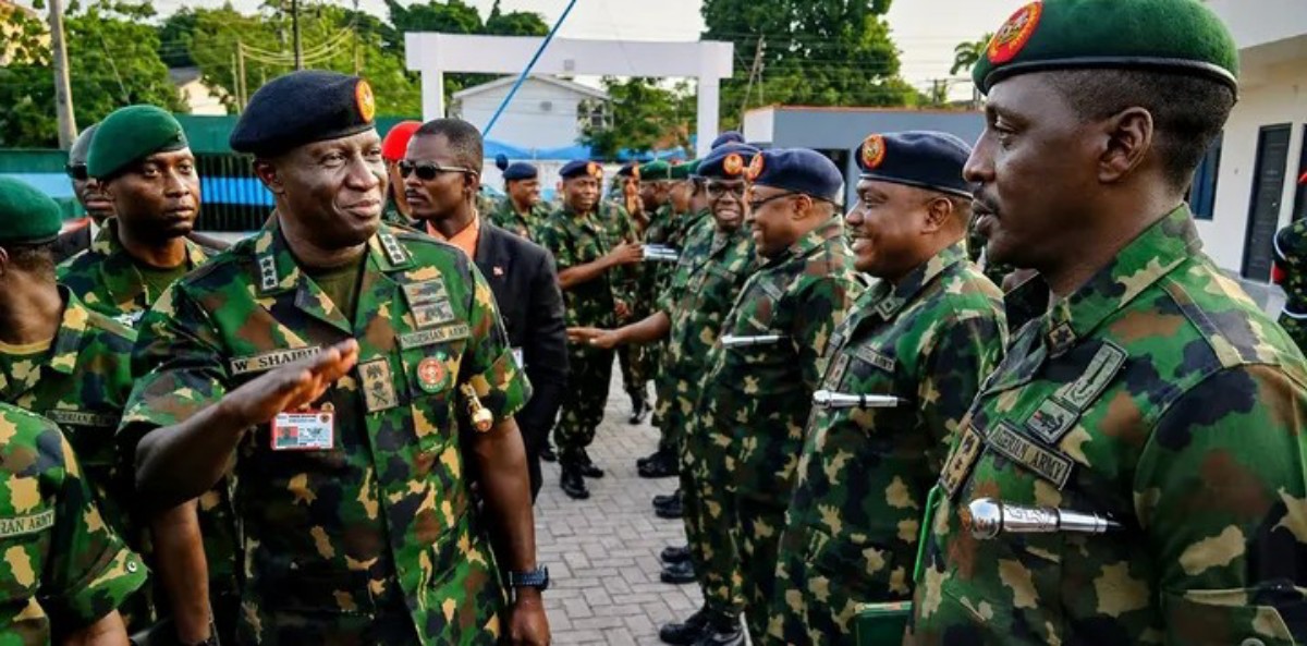 Senior army officer saluting as a line of soldiers in camouflage uniforms stand at attention outdoors, badges visible on uniforms.