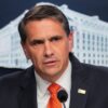 Man in a dark suit and orange tie speaks at a microphone during a press briefing, with a government backdrop behind him.