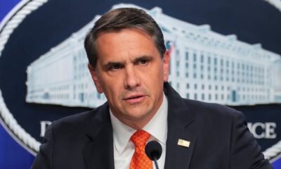 Man in a dark suit and orange tie speaks at a microphone during a press briefing, with a government backdrop behind him.