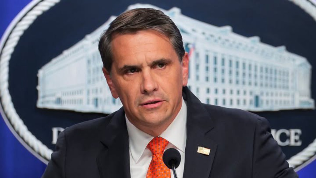 Man in a dark suit and orange tie speaks at a microphone during a press briefing, with a government backdrop behind him.